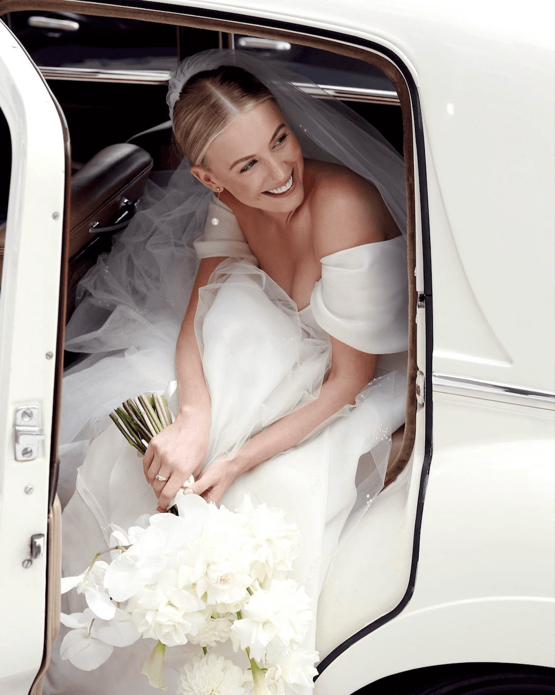A bride holding white flowers while seated in a vintage car, photographed by ImageHaus.