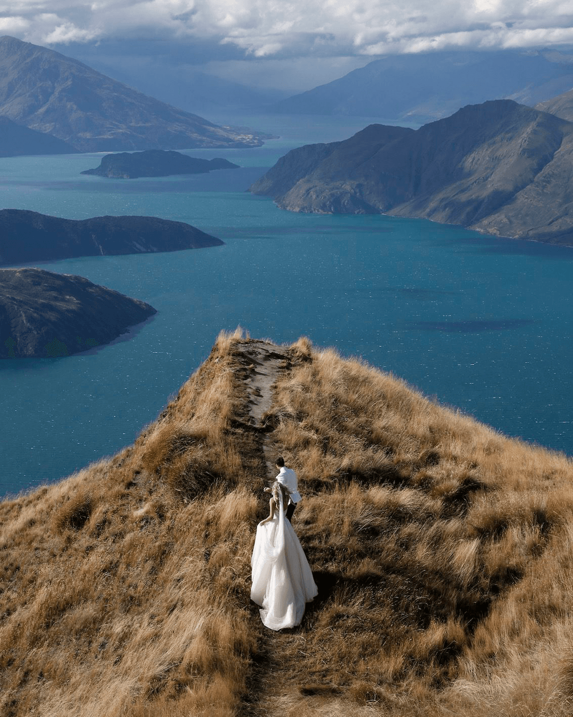 A wedding couple on a grassy mountain ridge above a lake, photographed by ImageHaus.